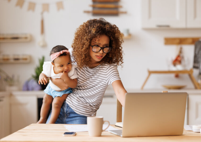 Multitasking young african american woman, working mother freelancer using laptop computer with baby girl in hands, ethnic female doing freelance work on maternity leave