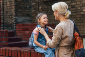 A tender moment of a mother supporting her cheerful daughter as she prepares for school. The setting portrays a warm bond and daily routine in a city environment, emphasizing education and parenthood.