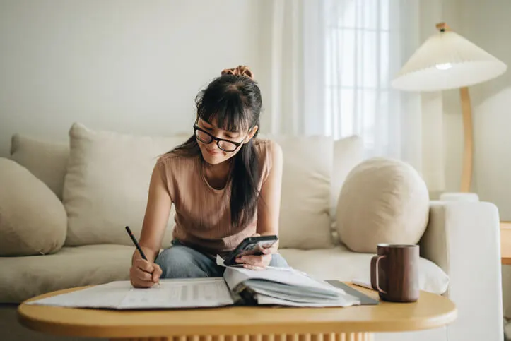 A woman sits at her living room with smartphone and financial reports doing her taxes.